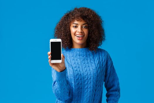 Look at my score. Happy and proud, good-looking african-american woman browsing dating app, showing smartphone screen camera, take a look, standing blue background in winter sweater.
