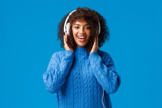 Happy charismatic african-american smiling girl got christmas present new headphones, listen music and enjoying awesome beats, touching earphones and looking camera, blue background.