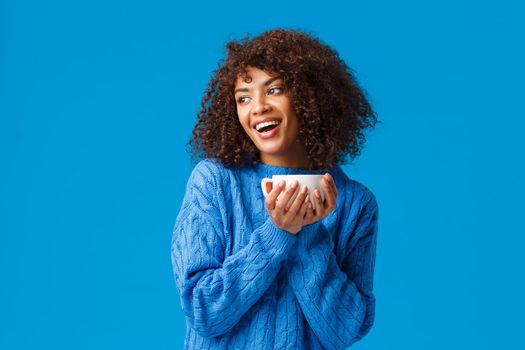 Girl having chat with girlfriend while drinking coffee. Cheerful and cute african-american lovely woman with afro haircut, in sweater, tilt head gazing left and holding hot cup delicious tea.