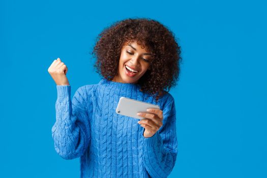 Excited happy african-american hipster girl in winter sweater, cheering raise fist up joyfully triumphing, winning level in game, holding smartphone horizontally and smiling.