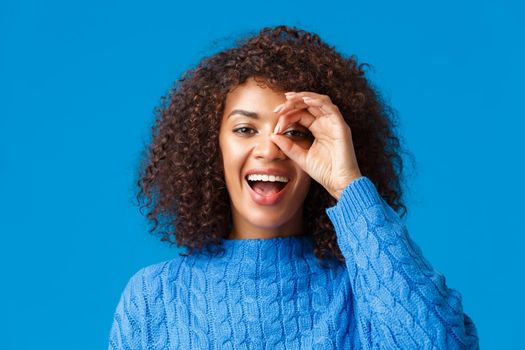 Looking into distance. Close-up cheerful attractive african-american woman searching something, found great holidays discount, look through okay sign and smiling pleased, blue background.