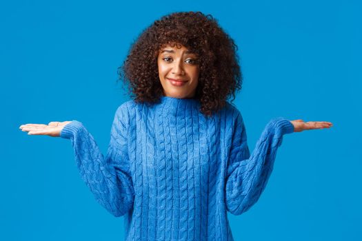 Who cares i dont know. Confused and clueless silly cute smiling african-american female in winter sweater, shrugging with hands spread sideways and smile as have no idea, standing blue background.