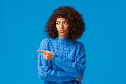 What a heck. Displeased and reluctant african-american bothered woman with afro haircut, pointing looking left with disapproval, express condemn or scorn, standing unsatisfied blue background.