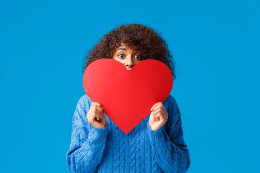 Shy and flushed cute girl scared confess to girl she loves her, holding big red hear and hiding face, peeking at camera with nervous scared gaze, standing blue background indecisive.