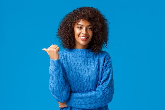 Happy friendly outgoing, pretty african-american female in winter sweater, sharing link, giving advice, pointing thumb left for you check-out advertisement, smiling standing blue background.