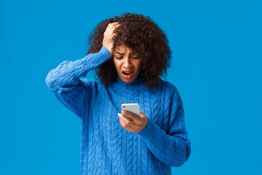 Troubled and upset, worried young woman feeling panic and anxiety, grab head, reading shocking news on smartphone, react to troublesome message, alarmed standing blue background.