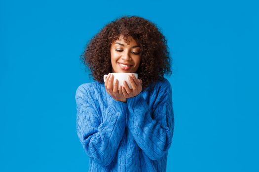 Coziness, home and holidays concept. Relaxed sensual and delighted, smiling tender african american woman with curly hair, looking at delicious warm tea, drinking coffee from cup, grinning.
