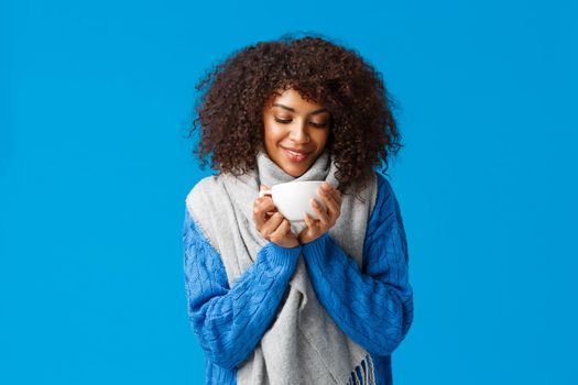 Tender and cute african-american woman with afro haircut, in sweater and scarf, looking at hot delicious cup tea, smiling and enjoying comfort and coziness of winter ski resort, blue background.