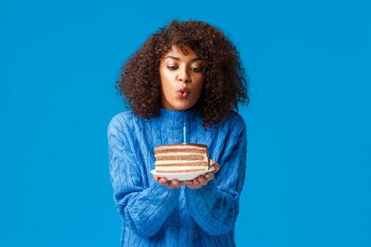 Cute and tender, young african-american woman with curly haircut, in sweater, holding b-day cake, blow-out candle and make wish to come true everything on birthday, standing over blue background.
