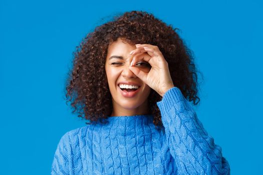 Carefree, happy good-looking african american woman with curly hair in sweater, looking through okay gesture, staring at something interesting from distance, standing blue background.