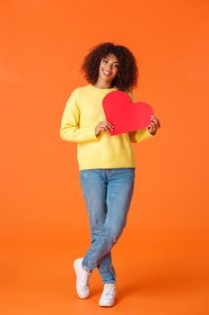 Full-length vertical shot attractive lovely smiling, romantic african-american girlfriend with afro, curly haircut, celebrating valentines day, holding big red cute heart and looking camera.