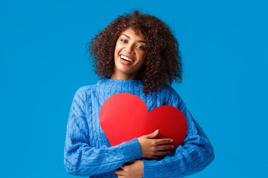 Cute and tender funny, smiling african-american female with afro haircut, press big red heart sign to chest and embrace it with delighted charming grin, showing love and affection, blue background.