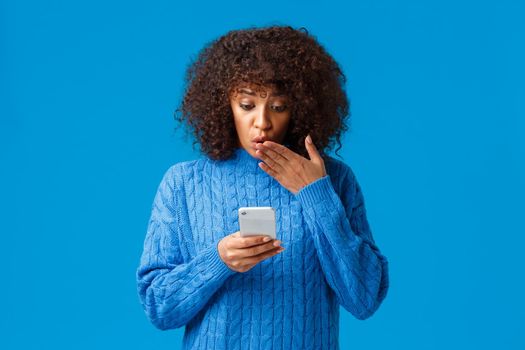 Oh gosh. Impressed and startled young african-american woman reading shocking news on internet, looking at smartphone display concerned, gasping cover mouth shook, blue background.