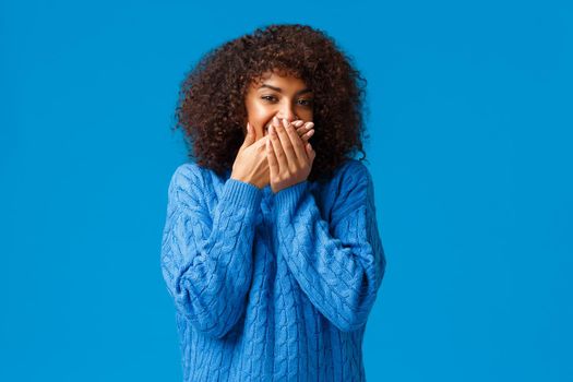 Cute and silly, lovely african-american woman with afro haircut, chuckling smiling and cover mouth as trying hold laugh, standing blue background making fun of friend, standing blue background.