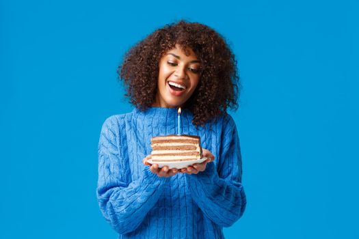 Celebration, holidays and party concept. Dreamy and lovely cute african-american woman with afro haircut, in sweater, tilt head and watching at lit candle on birthday cake, smiling making wish.