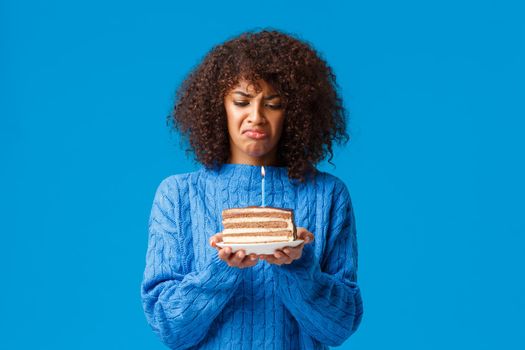 Upset and gloomy, distressed young african-american woman hate celebrating birthday feeling older, looking bothered and displeased at b-day cake with lit candle, sulking, blue background.