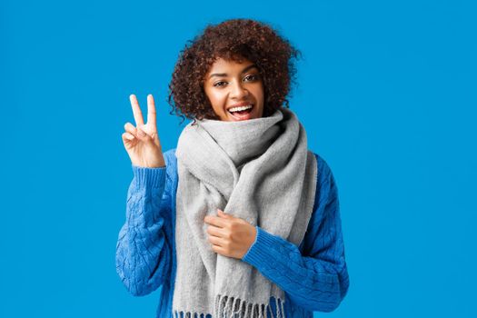 Cheerful cute african-american woman with afro haircut in winter sweater, warm comfortable scarf, showing peace sign and smiling, wishing happy holidays, best wishes, standing blue background.