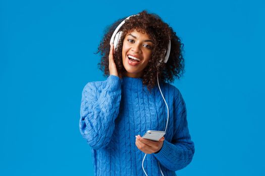 Happy and cheerful african-american woman playing karaoke game app in smartphone, wearing headphones singing and enjoying awesome music, sound quality, standing blue background.