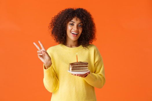 Waist-up portrait happy african-american woman in yellow sweater, showing peace sign and say cheese, birthday girl taking photo with b-day cake and candle, making wish, standing orange background.