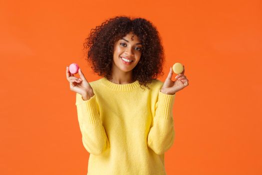 Waist-up portrait lovely young african-american female with curly afro haircut, holding two macarons and smiling, eating sweets, hate diets but like pastry and desserts, standing orange background.