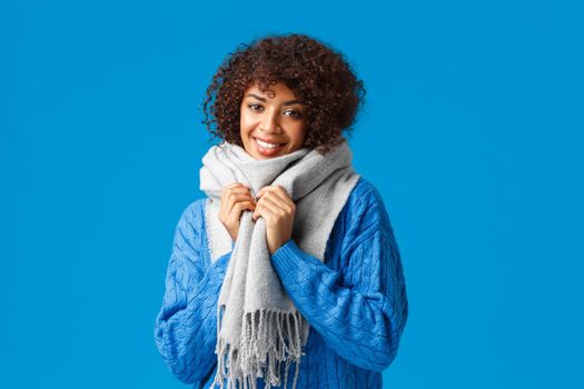 Waist-up shot charming smiling attractive african-american woman in winter sweater and warm scarf around neck, grinning and looking camera, wish happy holidays,s tanding blue background.