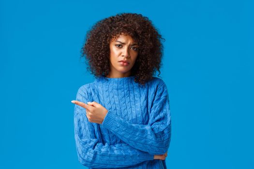 Explain to me, what is this. Displeased gloomy and unsatisfied african-american girlfriend with afro haircut, pointing finger left, frowning skeptical and disapproval, blue background.
