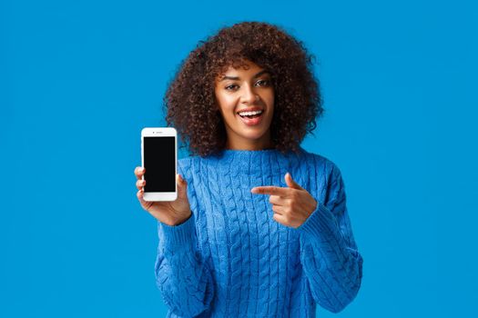 Check this out. Happy charismatic african-american woman with afro haircut, holding smartphone, showing mobile screen, pointing display as promote application, shopping app or game.