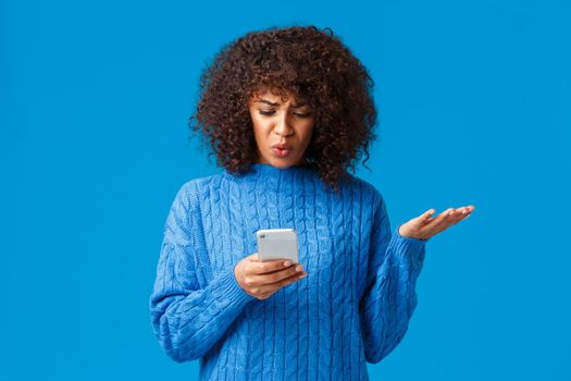 Problem, messaging and people concept. Confused and displeased, tensed african-american young woman receive strange message, staring at smartphone screen clueless, shrugging, blue background.