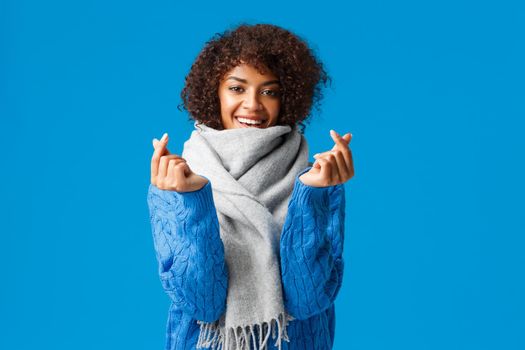 Romantic and silly cute african-american girlfriend, afro haircut, winter sweater and scarf, waiting valentines day present and date, showing korean heart signs and smiling, blue background.