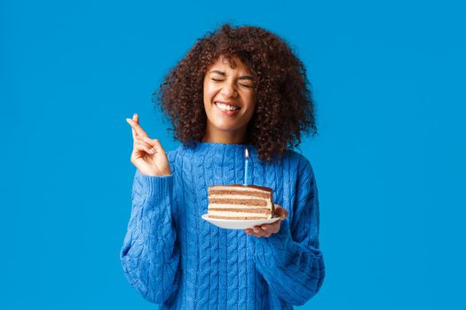 Hopeful and excited, cute african-american woman in sweater, making wish and cross fingers good luck, hope dream come true as close eyes and blowing-out candle on birthday cake.