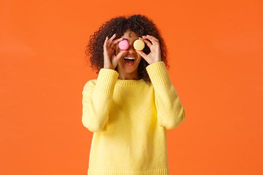 Playful funny and carefree, pretty african-american curly girl in yellow sweater, fool around make eyes from macarons and smiling, eating sweets, like desserts, standing orange background.