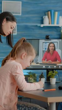 Pupil taking notes on textbook using video call with teacher while mother assisting online class lesson. Schoolgirl talking to professor on video conference for remote courses on computer.