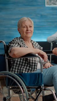 Portrait of old disabled woman and man nurse looking at camera in nursing home. Retired patient with chronic issues sitting in wheelchair while medical assistant preparing for examination