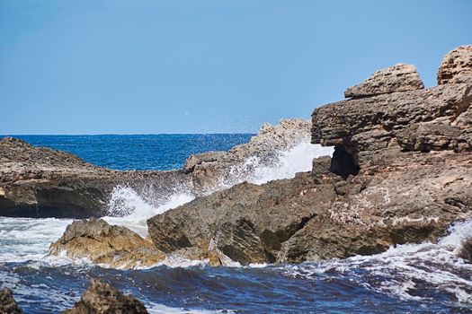 Rocks on the shore hit by the waves. Balearic Islands, Mediterranean Sea, blue sky, splashes of water.