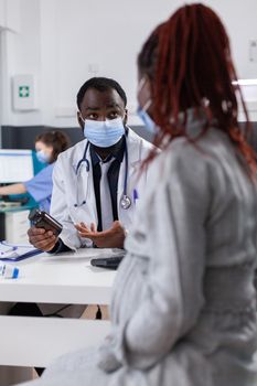 Doctor holding bottle of pills to pregnant patient in medical cabinet. Patient expecting baby receiving flask with prescription medicine and treatment from doctor, wearing face masks