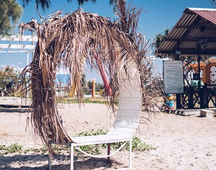 Real nature beauty background. Sand beach park place for romantic relax time. White wooden round bench, dry palm leaves shade beige canopy. Turquoise blue sky. Unusual trendy modern outdoor furniture.