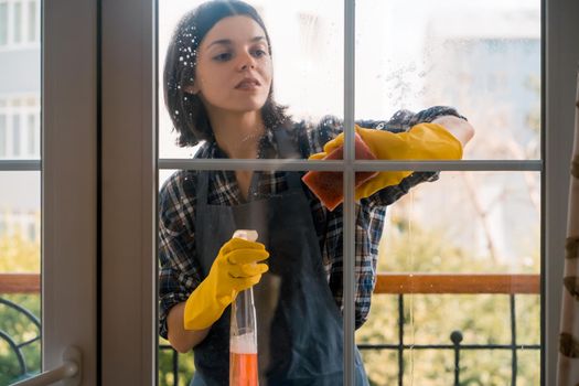 A young girl in yellow rubber gloves and a uniform washes the windows thoroughly with a detergent, wiping them dry with a rag, the woman is cleaning the house.