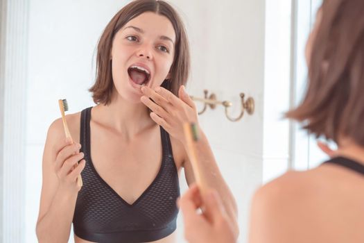 A young smiling girl brushes her teeth with toothpaste in the bathroom by the mirror in the morning, a woman takes good care of the health of tooth enamel and gums.