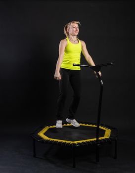 Girl on a fitness trampoline on a black background in a yellow t-shirt black fit sport, equipment body caucasian healthy muscular bounce. Wellness white trainer, sportswear muscle instructor enjoy