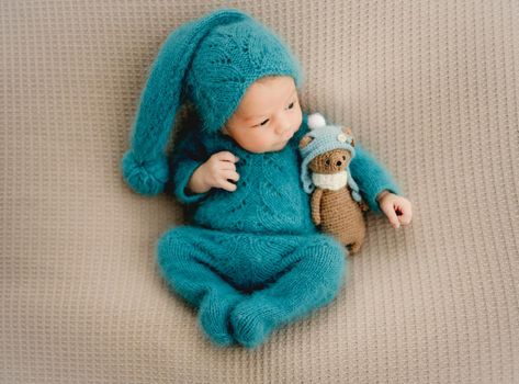 Newborn baby boy wearing knitted costume and hat holding teddy bear toy and looking back. Cute infant child kid studio portrait