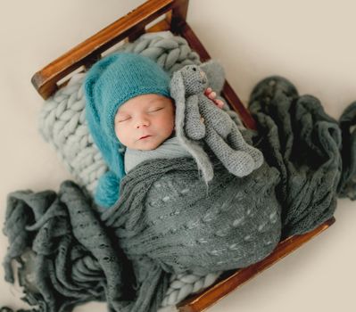 Newborn baby boy wearing knitted hat sleeping in tiny bed and holding bunny toy. Cute infant child kid studio portrait