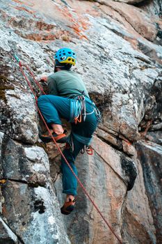 The young man is engaged in extreme sports, leads an active lifestyle, climbs a mountain, a rock with climbing equipment, wearing a helmet, with a rope, carabiners and climbing shoes.