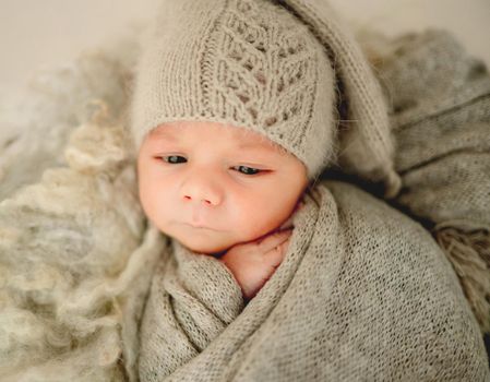 Newborn baby boyvswaddled in knitted fabricand wearing hat falling asleep. Cute infant child kid closeup studio portrait