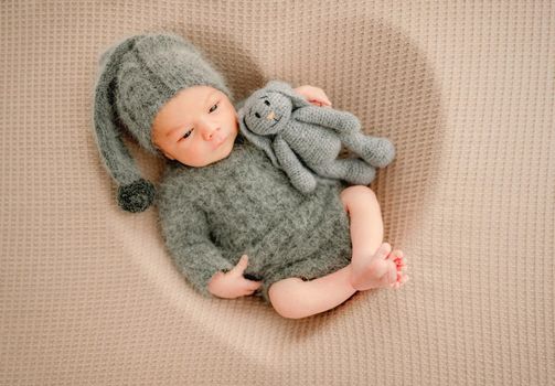 Newborn baby boy wearing knitted costume and hat sleeping in heart shape and holding bunny toy. Cute infant child kid studio portrait
