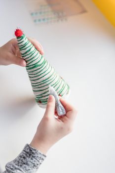 Kid is holding a Christmas tree decoration in his hands. Kid is showing a Christmas tree decoration. Merry Christmas tree project for kids. Scissors, green cotton yarn on a wooden table