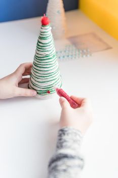 Kid is holding a Christmas tree decoration in his hands. Kid is showing a Christmas tree decoration. Merry Christmas tree project for kids. Scissors, green cotton yarn on a wooden table