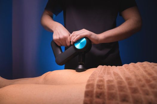 Close-up of a professional male masseur stimulates the leg muscles of a male patient in a dark spa room for massage. Percussion mechanical effect on overstrained muscles.