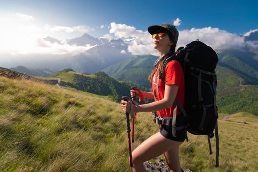 A tourist girl with a backpack admires the sunset from the mountainside. Traveler in the background of mountains and clouds at sunset. Woman tourist in sunglasses, cap and with a backpack stands on the slope of the mountain. Trekking and hiking in the mountains