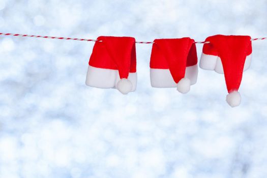 Three red little Christmas hats hang from a rope against a blurred shiny blue-gray background. Copy space, concept.