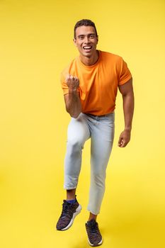 Vertical full-length cheerful sportsman in casual orange t-shirt, white pants, satisfied with excellent results, fist pump and smiling say yes as winning, achieve goal or prize, become champion.